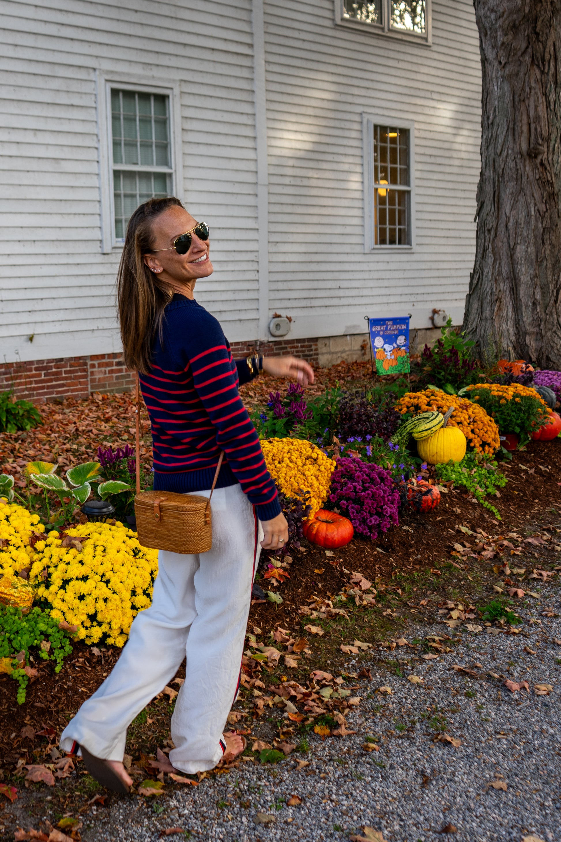 Woman walking in a garden with colorful flowers and pumpkins, wearing sunglasses and a striped shirt.