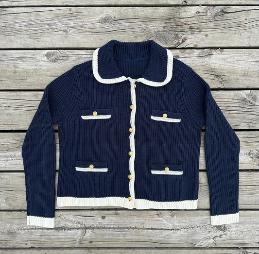 A navy blue cardigan with ivory collar, cuffs, and hem, featuring nautical buttons and two front welt pockets.