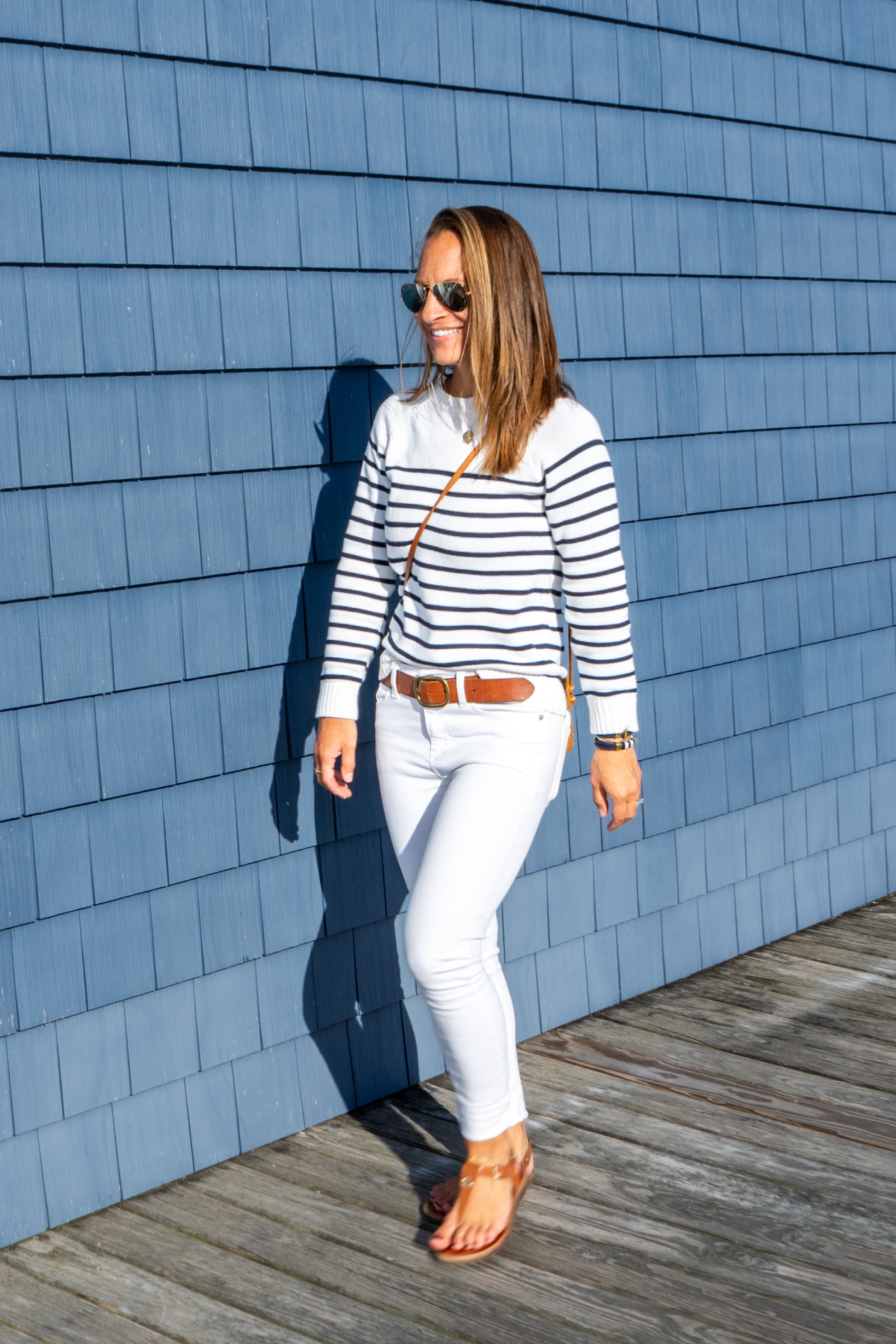 Woman in a striped sweater and white pants standing against a blue shingled wall.