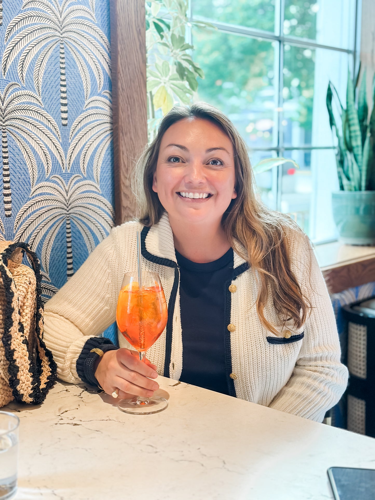 Woman holding an aperol spritz in a cafe with palm tree wallpaper.